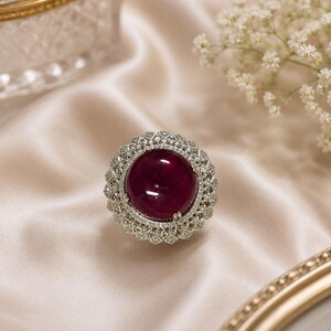 May include: A silver ring with a large, round, deep red gemstone at the center, surrounded by a halo of smaller, sparkling stones. The ring is set against a backdrop of cream-colored satin.