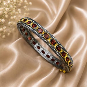 May include: A silver-toned bangle bracelet with oval, deep red gemstones set in gold-colored bezels. The bracelet is accented with small, clear stones and rests on a satin surface.