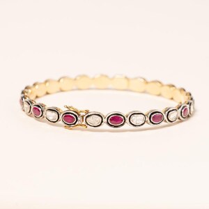 Puede incluir: Una pulsera de oro y plata con piedras preciosas rojas y claras de forma ovalada. La pulsera presenta un cierre de oro y una banda de oro en el interior. Las piedras están engastadas en un bisel de plata.