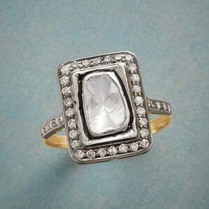 May include: A rectangular ring featuring a large, clear, faceted stone at the centre, encircled by smaller, clear stones. The ring has a gold band and a silver-coloured setting around the central stone and the smaller stones.
