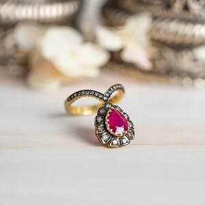 May include: A gold ring with a teardrop-shaped, bright red gemstone. The gemstone is surrounded by smaller, clear stones. The ring band is also adorned with small, clear stones. The ring is set against a soft, blurred background.
