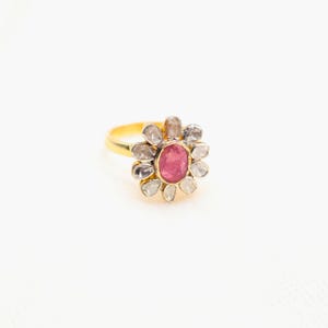 Può includere: Un anello d'oro con una gemma rosa ovale centrale, circondata da un alone di gemme più piccole e trasparenti. Il design dell'anello ricorda un fiore, con le gemme disposte a petali. L'anello è posto su uno sfondo bianco.