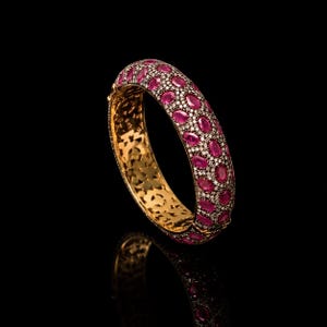 Op de afbeelding: Een gouden armband met een dichte schikking van ovaal geslepen rozerode robijnen en kleine witte pavé gezette diamanten. Het buitenoppervlak is volledig ingelegd met deze edelstenen, terwijl de binnenband een ingewikkeld uitgesneden patroon vertoont.