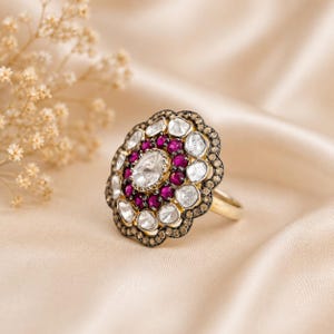 Può includere: Un anello d'oro con un design floreale. L'anello presenta una grande pietra trasparente di forma ovale al centro, circondata da pietre più piccole, rotonde, rosse e trasparenti. L'incastonatura dell'anello è marrone scuro e la fascia è dorata.