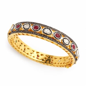 Może przedstawiać: Złota bransoletka typu bangle z zawiasem, ozdobiona rzędem naprzemiennie ułożonych okrągłych czerwonych rubinów i białych diamentów w ciemnych oprawach. Zewnętrzna obręcz jest wysadzana małymi czarnymi diamentami, a wewnętrzna struktura ma ażurowy wzór.