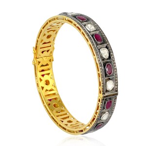Puede incluir: Una pulsera de brazalete dorada con un cierre de bisagra. La pulsera presenta una fila de piedras preciosas rojas y blancas alternas engastadas en una banda de metal oscuro. La pulsera tiene un diseño calado.