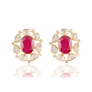 May include: A pair of gold-toned stud earrings. Each earring features a large, oval-shaped, deep red ruby at the center, surrounded by a halo of irregularly shaped, clear gemstones. Small, round, clear gemstones are interspersed around the halo.