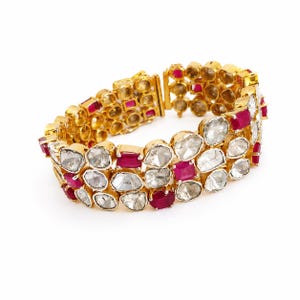 Puede incluir: Una pulsera de oro con tres filas de piedras blancas translúcidas irregulares y piedras rojas rectangulares vibrantes. La pieza está construida con un marco de metal articulado, mostrando un patrón repetitivo de estas gemas en oro amarillo.