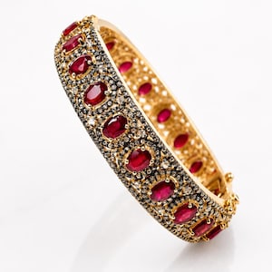 May include: A gold hinged bangle bracelet featuring a row of oval-cut deep red rubies set against a dark, textured background encrusted with numerous small, clear, sparkling stones. The interior of the bracelet has a polished gold finish.