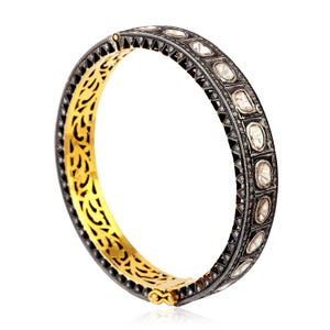 Könnte beinhalten: Ein aufklappbares Armreif-Armband mit goldener Innenseite und silberner Außenseite. Die silberne Außenseite ist mit quadratisch gefassten, klaren Edelsteinen besetzt. Die goldene Innenseite hat ein dekoratives, durchbrochenes Design. Das Armband wird vor weißem Hintergrund gezeigt.