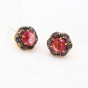 May include: A pair of gold-toned stud earrings. Each earring features a round, pink gemstone surrounded by a halo of small, dark-colored stones. The earrings are set against a white background.