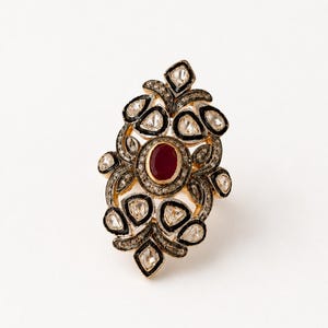Puede incluir: Un anillo de oro con una piedra preciosa roja de corte ovalado en el centro, rodeada por múltiples piedras claras de forma irregular. El engaste incluye detalles en esmalte negro y un acabado de metal oscuro texturizado alrededor de las piedras, creando un diseño floral alargado.