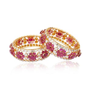 May include: Two gold bangles with intricate floral designs. The bangles are adorned with vibrant red gemstones and sparkling white stones, creating a luxurious and eye-catching accessory. The jewelry is set against a clean white background.
