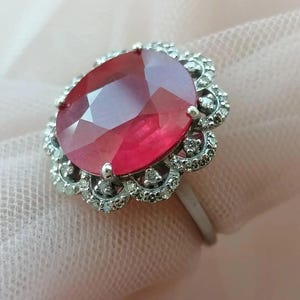 May include: A silver ring with a large, oval-cut ruby. The ruby is a deep red hue and is encircled by smaller, clear gemstones. The ring's band is a simple, polished silver. The ring's design is classic and elegant.