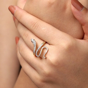 May include: A silver and gold snake ring with a coiled design. The snake's head is adorned with a red stone and the body is encrusted with small, clear stones. The ring is worn on a finger.