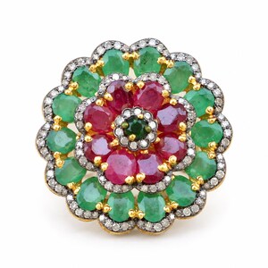 May include: A floral-shaped ring featuring a central dark green gemstone surrounded by a layer of oval-cut red rubies. An outer ring of oval-cut green emeralds frames the design, with small pavé-set diamonds lining the scalloped edges in gold.