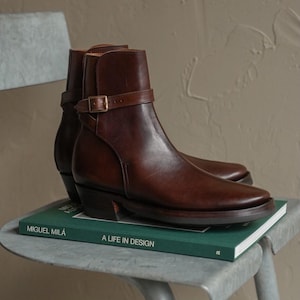 May include: A pair of dark brown leather ankle boots featuring a slim strap with a metal buckle around the ankle. The boots have a smooth finish, a rounded toe, and a stacked heel. The boots are resting on a green book titled Miguel Milá A Life in Design.