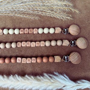 May include: Three personalised baby dummy clips with wooden beads spelling "MILA", "EVELYN", and "NOAH". Each clip has a wooden round clip and a loop. The clips are displayed on a brown surface with decorative dried plants.