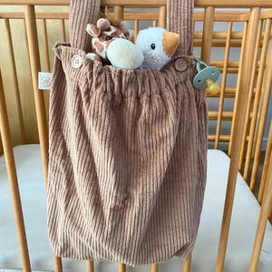 Könnte beinhalten: Ein hellbrauner Babybett-Organizer aus Cord, der an einem Holzbett hängt. Der Organizer enthält Stofftiere und einen Schnuller. Der Organizer hat einen Knopfverschluss.