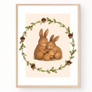 Op de afbeelding: Een ingelijste prent met een aquarel illustratie van een konijnenfamilie. Twee volwassen konijnen omarmen een babykonijn, alles binnen een groene en eikelkrans. De achtergrond is zacht perzik van kleur.