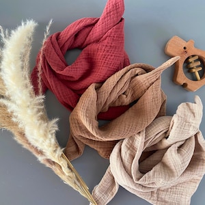 Könnte beinhalten: Drei Baby-Lätzchen aus Musselin in Rot-, Braun- und Beigetönen sind auf einer grauen Oberfläche angeordnet. Ein hölzernes Babyrassel in Form eines Nilpferds und ein Bündel getrockneter Pampasgras sind ebenfalls im Bild. Die Lätzchen sind weich und strukturiert.