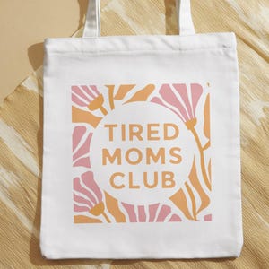Op de afbeelding: Witte draagtas met een bloemenontwerp in roze en oranje. De woorden "TIRED MOMS CLUB" staan in een cirkel in het midden van de tas. De tas heeft witte handvatten en ligt op een beige ondergrond.