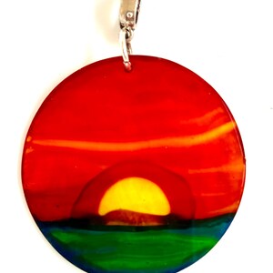 Könnte beinhalten: Ein runder Anhänger mit einem Sonnenuntergangsdesign. Der Anhänger zeigt einen leuchtend roten Himmel, eine gelbe Sonne, die über einem dunklen Berg untergeht, und einen grünen und blauen Ozean. Er hat eine silberfarbene Öse zum Anbringen an einer Kette.