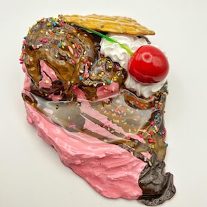 May include: A whimsical sculpture of a sundae slice, featuring pink ice cream, chocolate sauce, whipped cream, a cherry, and a waffle. The sundae is decorated with colorful sprinkles, creating a playful and vibrant aesthetic.