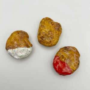 May include: Three decorative, textured stones with a golden-brown hue. One stone has a white and green base, another has a red base, and the third is plain. The stones are arranged on a white surface.