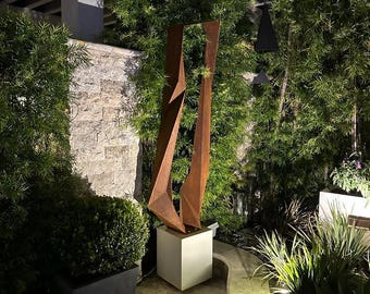 Könnte beinhalten: Eine moderne Außenskulptur mit geometrischem Design. Die Skulptur aus verwittertem Metall steht auf einem weißen Würfelsockel. Das Kunstwerk befindet sich in einem Garten mit üppigem Grün und Steinwegen.