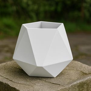Puede incluir: Una maceta geométrica blanca con un diseño multifacético. La maceta tiene una parte superior pentagonal y una base en forma de diamante. Está sobre una superficie de piedra gris desgastada, con un fondo verde borroso.