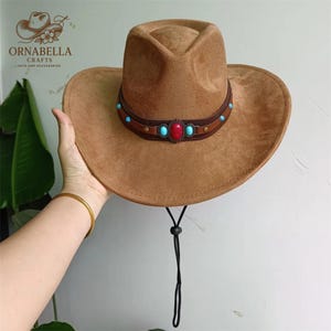 May include: A brown cowboy hat with a decorative band featuring turquoise and red stones. The hat has a wide brim and a black chin strap. The hat is a suede-like material.