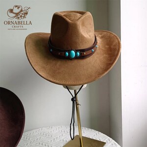 May include: A brown cowboy hat with a wide brim and a decorative leather band. The band is adorned with turquoise-colored stones. The hat is displayed on a gold-colored stand. The logo "ORNABELLA CRAFTS" is visible.