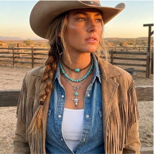 May include: A woman wearing a brown suede fringe jacket, denim shirt, and cowboy hat. She is wearing three turquoise necklaces, including a choker, a beaded necklace, and a pendant necklace with a bull skull charm.