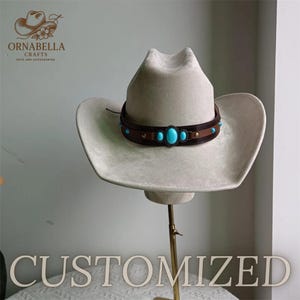Op de afbeelding: Een lichtgrijze cowboyhoed met een bruine leren band versierd met turkooizen stenen. De hoed wordt op een mannequinhoofd getoond. Het woord "CUSTOMIZED" staat onderaan de afbeelding.