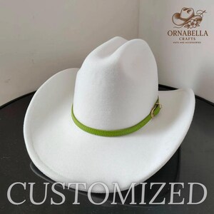 May include: A white cowboy hat with a green band and gold-colored buckle. The hat has a wide brim and a rounded crown. The word "CUSTOMIZED" is printed at the bottom of the image. The logo for Ornabella Crafts is in the upper right corner.