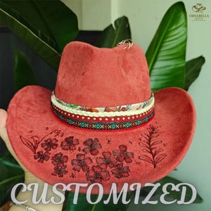 Peut inclure: Un chapeau de cowboy de couleur corail avec une bande décorative et un motif floral. Le chapeau présente une bande à motifs avec des accents rouges, blancs et bleus. Le mot "CUSTOMIZED" est imprimé sur le bord.