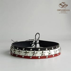 Könnte beinhalten: Ein schwarzes, weißes und rotes Choker-Halsband mit silbernen Sternen und Nieten. Das Halsband hat einen schwarzen Kordelverschluss. Das Markenlogo "ORNABELLA CRAFTS" ist oben rechts sichtbar. Ein modisches Accessoire.