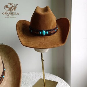 May include: A brown cowboy hat with a wide brim and a decorative band featuring turquoise-colored stones. The hat is displayed on a gold-colored stand. The logo "ORNABELLA CRAFTS HATS AND ACCESSORIES" is visible in the upper left corner.