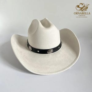 May include: A cream-colored cowboy hat with a black band adorned with silver-toned embellishments. The hat has a classic western design with a wide brim and a creased crown. The hat is a stylish accessory.