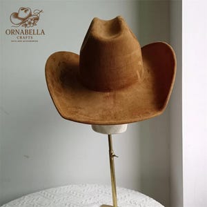 May include: A brown suede cowboy hat with a wide brim and a pinched crown. The hat is displayed on a hat stand with a gold-colored metal pole. The hat has a classic western style and is likely made for outdoor wear.