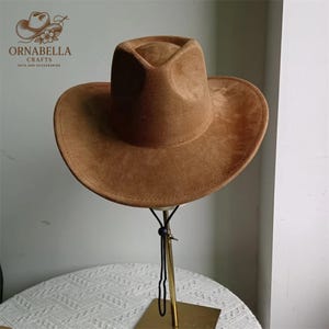 May include: A brown suede cowboy hat with a wide brim and a structured crown. The hat is displayed on a gold-colored stand. The Ornabella Crafts logo is visible in the upper left corner.