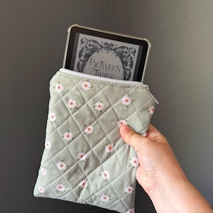 Könnte beinhalten: Eine gesteppte Stoffhülle in Salbeigrün mit weißem Gänseblümchenmuster und weißem Reißverschluss. Die Hülle enthält ein Tablet, auf dessen Bildschirm der Text Between Tides zu sehen ist. Die Steppung weist ein durchgehendes Rautenmuster auf.