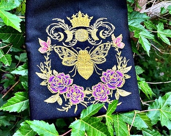 May include: A black fabric pouch with a gold embroidered design featuring a bee with a crown, surrounded by flowers and leaves.