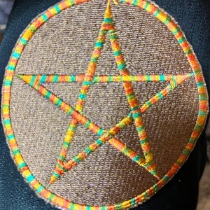 Op de afbeelding: Een bruine, geborduurde patch met een vijfpuntige ster in het midden. De ster is omrand met oranje, groen en geel garen. De patch is omgeven door een cirkel van oranje, groen en geel garen.