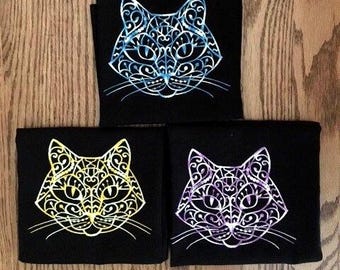May include: Three black t-shirts, each featuring a stylized cat head graphic. The top cat is blue, the bottom left cat is yellow, and the bottom right cat is purple. Each cat design is composed of intricate, swirling white line patterns.