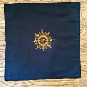 Op de afbeelding: Een zwart vierkant kussen met een gouden mandala-ontwerp van een zon met stralen.