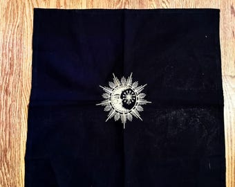Può includere: Un panno quadrato nero con un elaborato disegno argentato di sole e luna al centro. Il sole ha raggi appuntiti e la luna ha una faccia. Il panno è piegato ordinatamente su una superficie di legno.