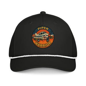 May include: Black baseball cap with a white rope detail and a circular patch. The patch features a cartoon airplane with the text "PIPER WARRIOR" in orange and blue. The airplane has a propeller and a menacing expression.