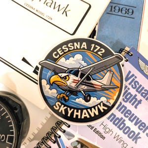 May include: A collection of aviation-themed items, including a Cessna 172 Skyhawk sticker with an eagle mascot, a notebook, and manuals. The sticker features a cartoon airplane design with the text "Cessna 172 Skyhawk."